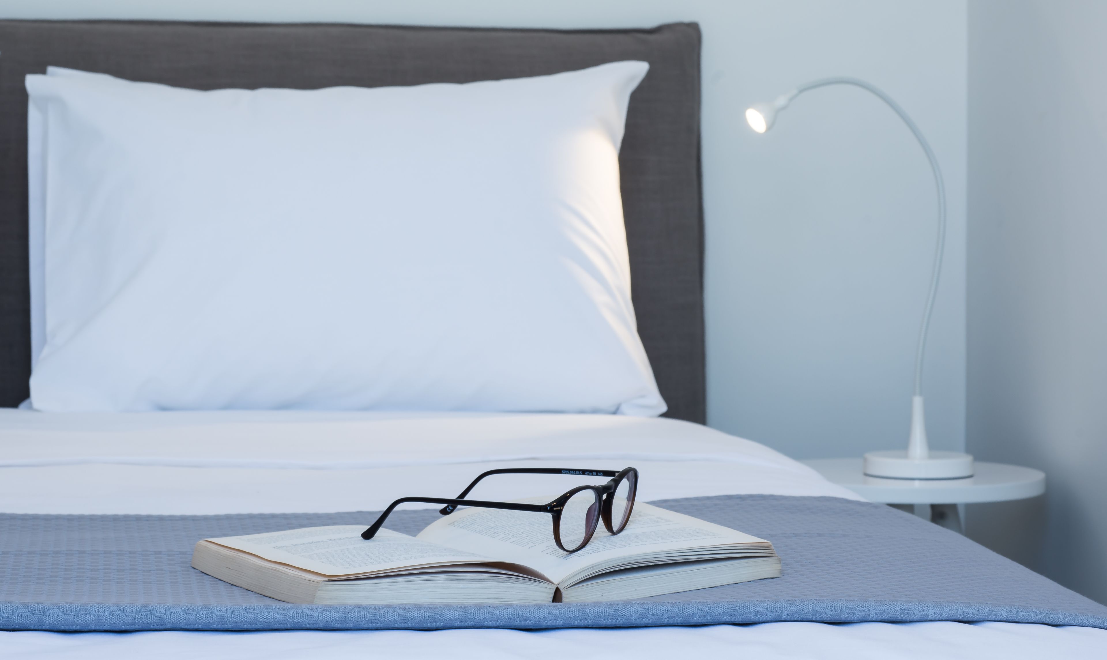 Una cama bien hecha con un almohadón blanco y un cabecero gris, un libro abierto con gafas encima, y una mesita de noche con una lámpara blanca.