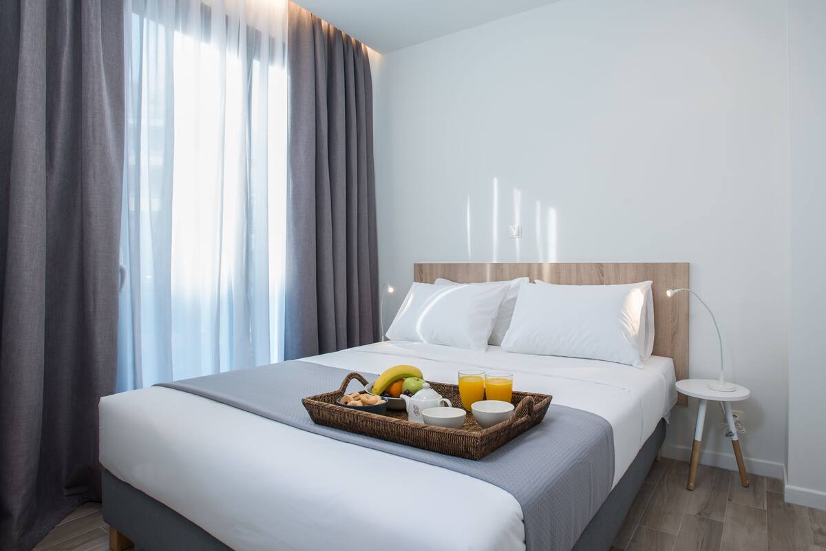A hotel room with a bed, a bedside table, a lamp, and a breakfast tray with fruits and drinks.