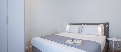 A neatly made bed with a grey and white duvet, a bedside table with a lamp, and a wardrobe.