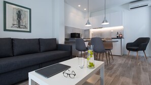 A modern living room with a grey sofa, a dining table with a laptop and glasses, and a kitchen area with a microwave and chairs. - Spot Apart (Athens)