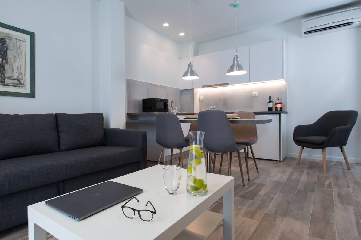 A modern living room with a grey sofa, a dining table with a laptop and glasses, and a kitchen area with a microwave and chairs.