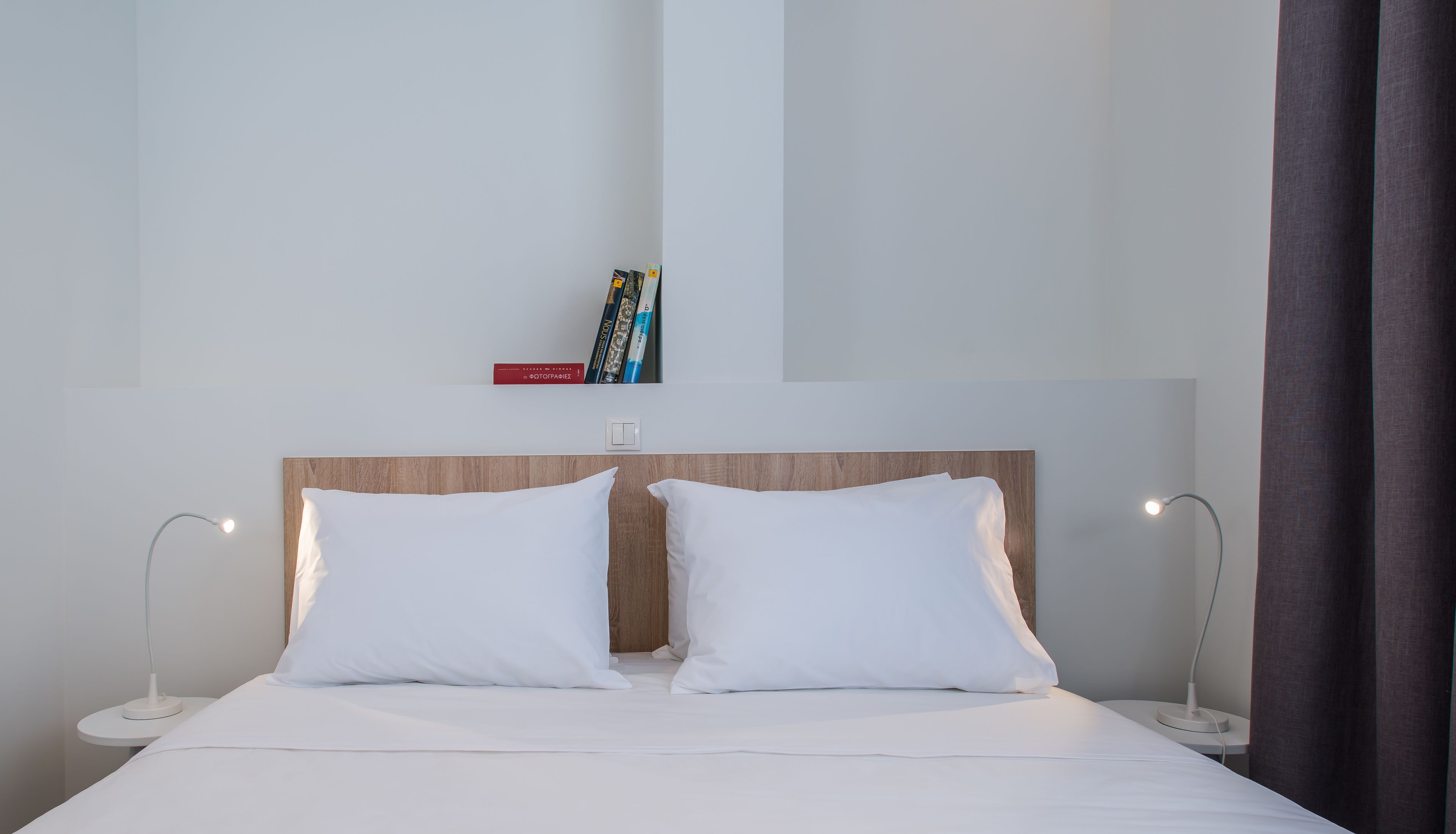 Une chambre d’hôtel avec une tête de lit en bois, deux oreillers blancs, une couvre-lit blanche, une table de chevet avec une lampe et une étagère avec des livres.