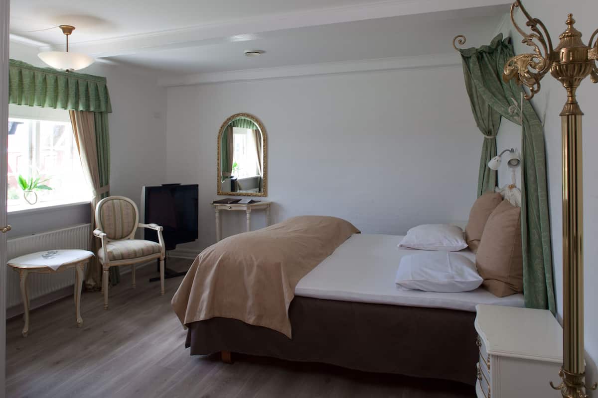 A bedroom with a bed, a chair, a desk with a television, a mirror, and a window with curtains.