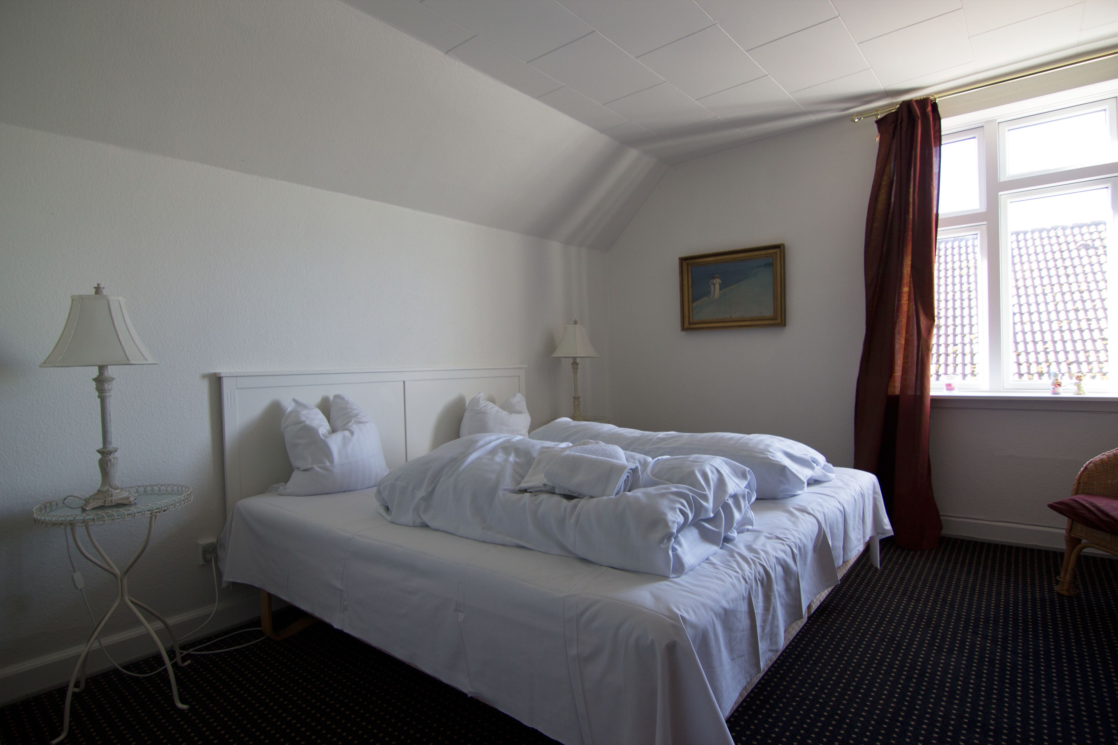 A hotel room with a neatly made bed, a small table with a lamp, a window with curtains, and a painting on the wall.