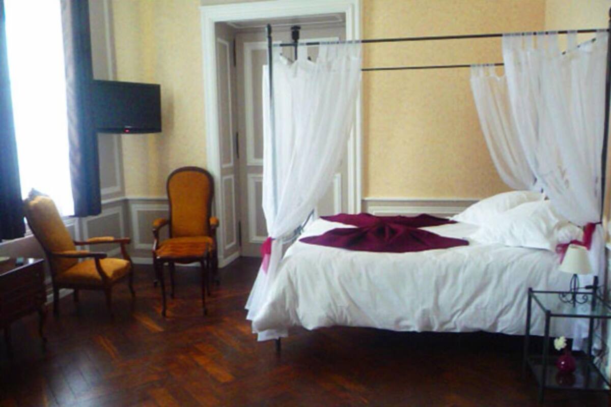 Quarto com cama de dossel, uma cadeira, mesa de cabeceira e televisão fixada na parede.