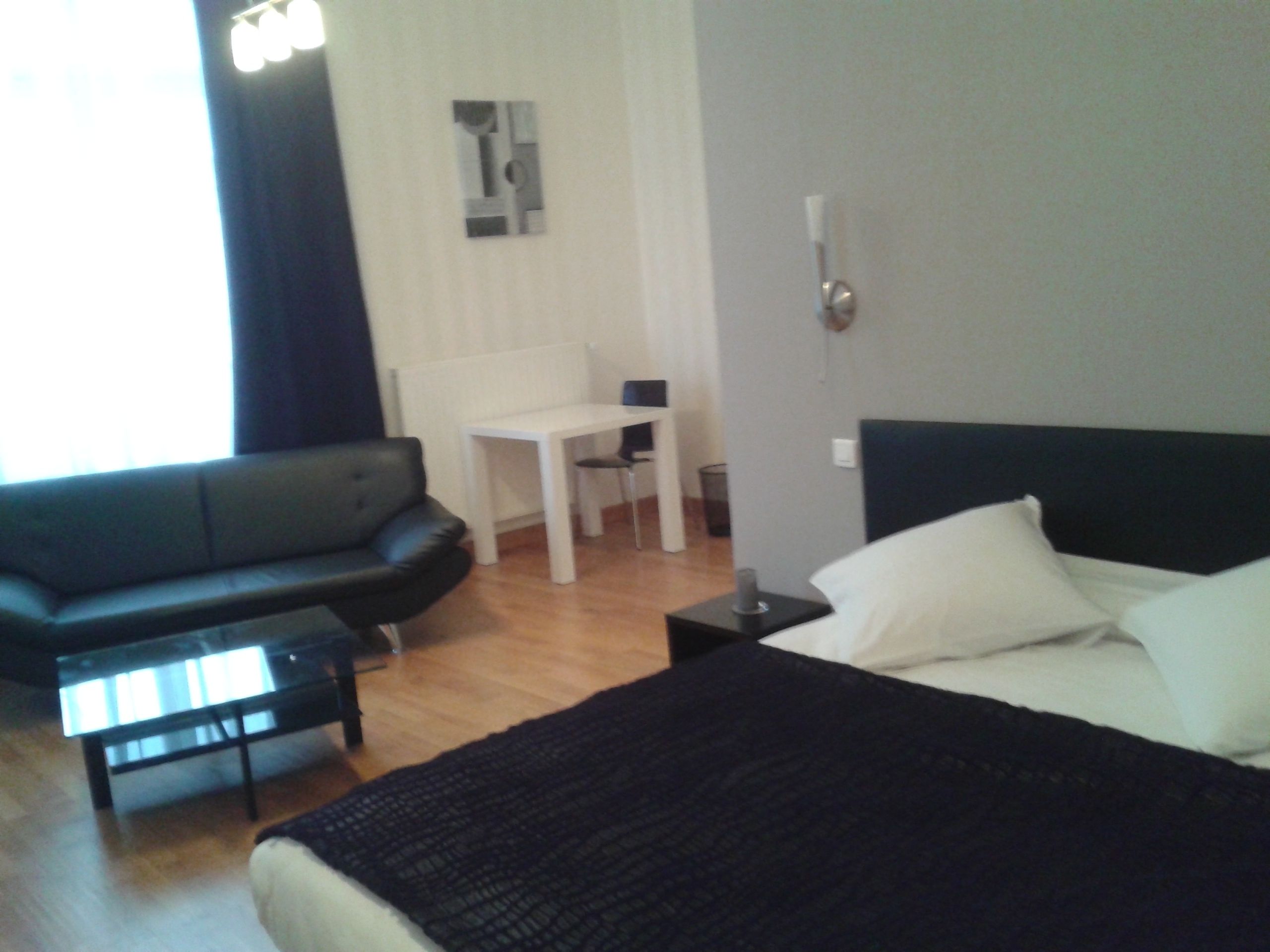 Una habitación de hotel moderna con un sofá de cuero negro, un escritorio blanco, una mesita pequeña, una cama con almohadas blancas y un aplique de pared.
