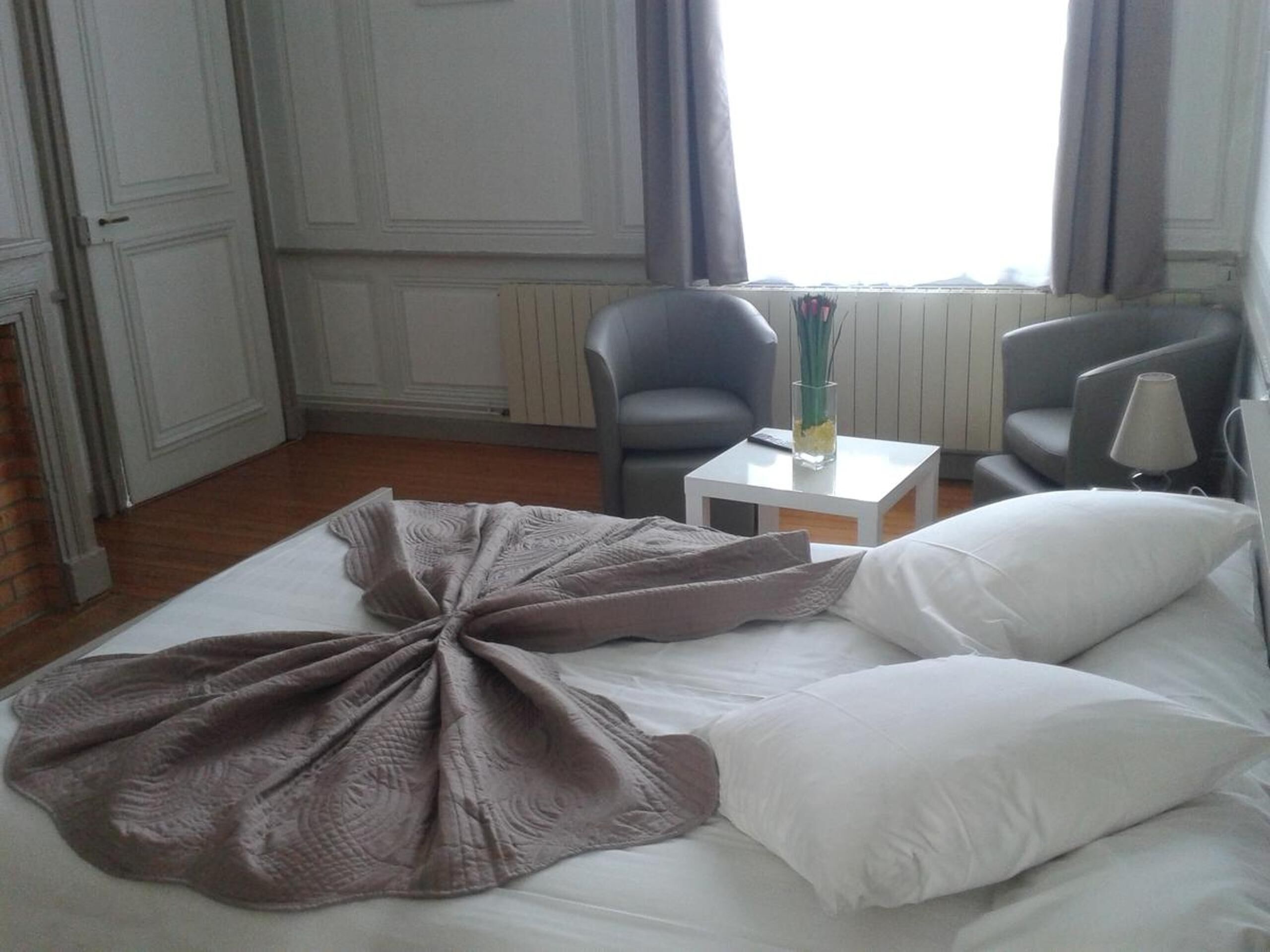 A neatly made bed with a decorative blanket, two grey armchairs, a small white table with a vase, and a lamp.