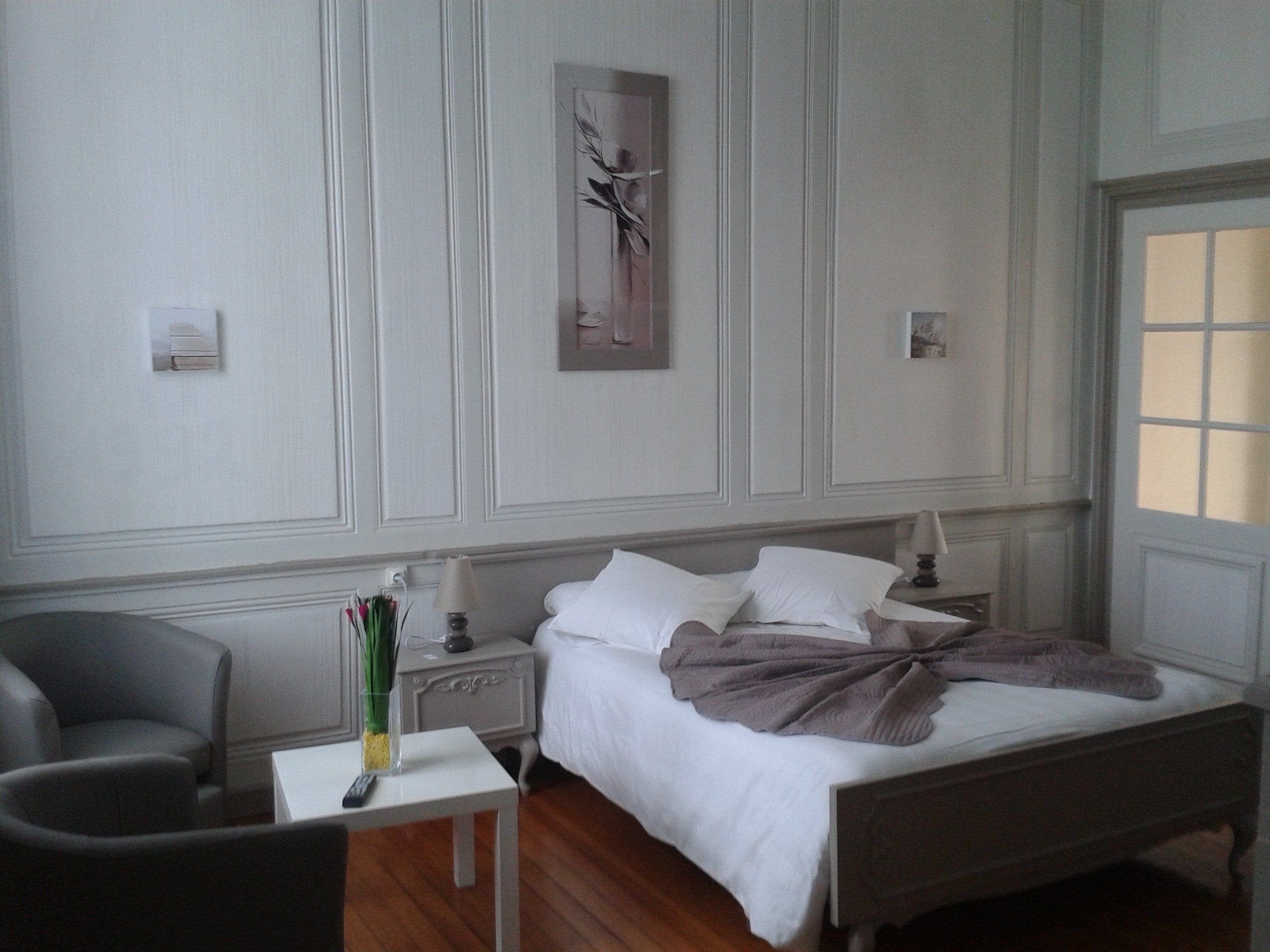 Een hotelkamer met een bed, nachtkastje, lamp, stoel en een schilderij aan de muur.