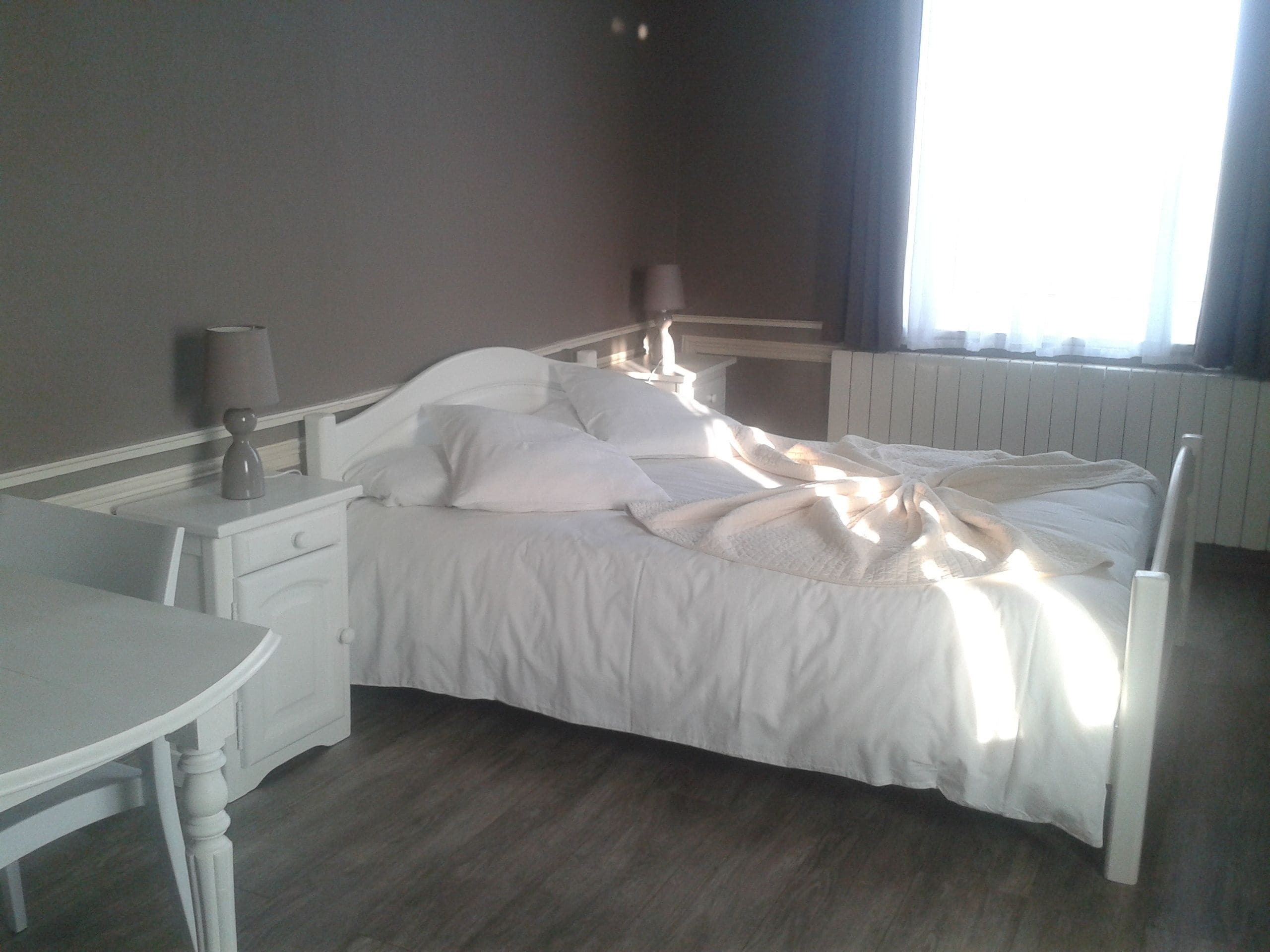Ein ordentlich gemachtes Bett mit weißen Leinen und Kissen, ein Nachttisch mit Lampe und ein Fenster mit Vorhängen.