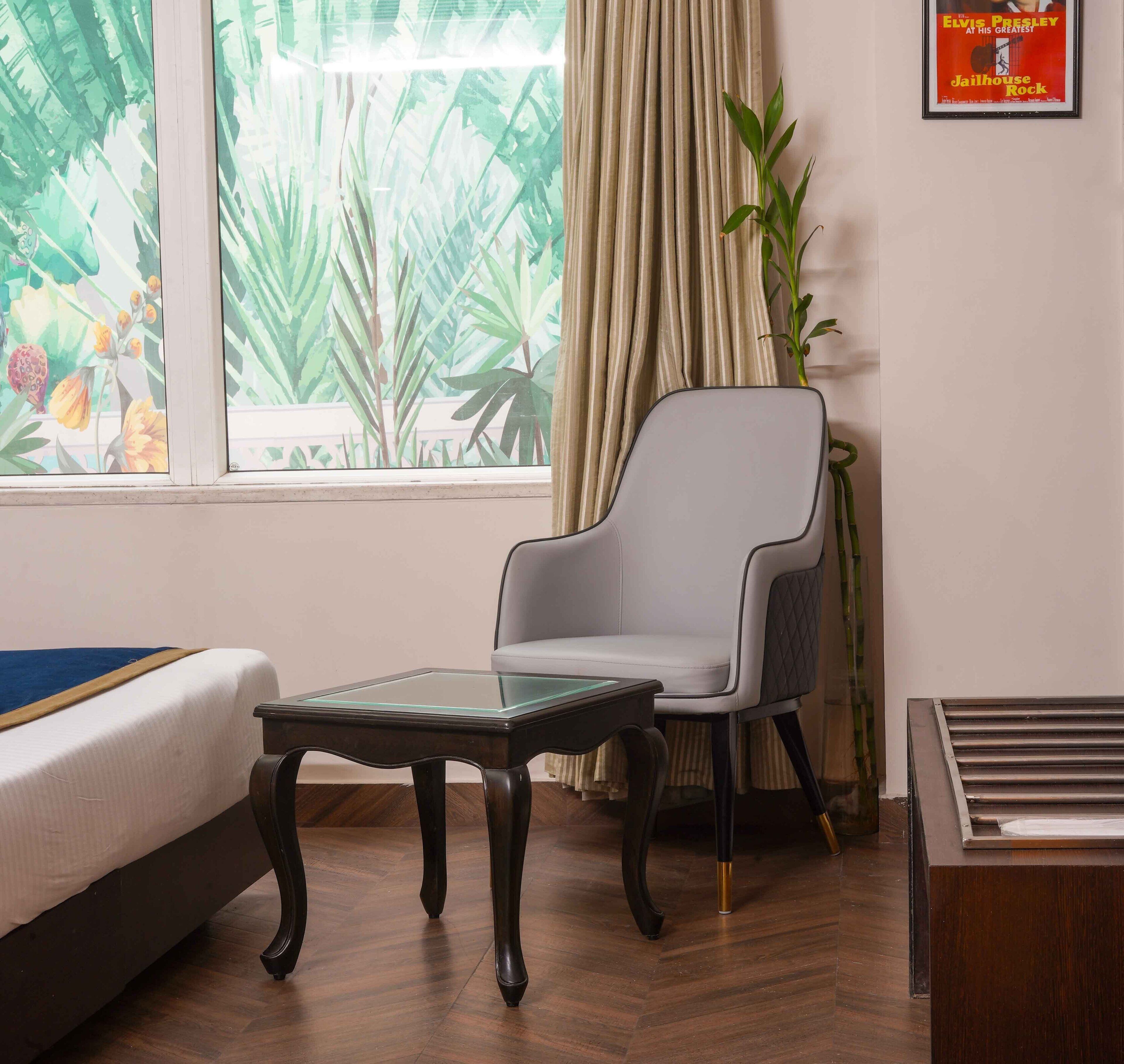 Habitació d'hotel amb un llit, una taula de vidre, un sofà armchair gris, una planta en test i un cartell a la paret.