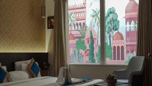 A hotel room with a bed, a chair, a small table with a plant, and a window with a view of a building. - Woodapple Residency (New Delhi)