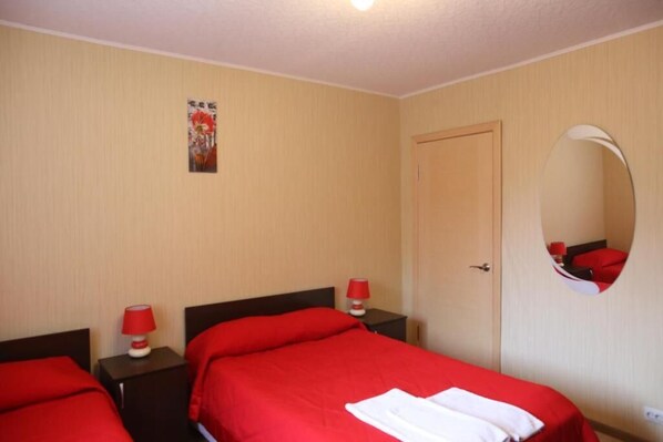 Habitación de hotel con dos camas, cada una con ropa de cama roja, una puerta de madera, una mesita de noche pequeña con una lámpara roja y un espejo montado en la pared.