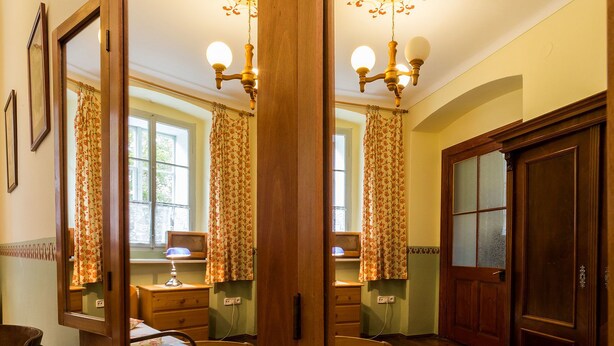 A traditional room with a wooden floor, a large mirror, a wooden wardrobe, a desk with a lamp, and a window with curtains. - Pension Gardena Kaplická 21 (Cesky Krumlov)