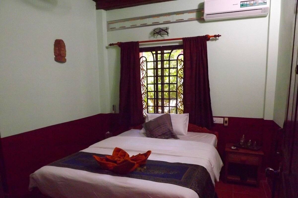Una habitación de hotel con cama, mesita de noche, ventana con cortinas y un equipo de aire acondicionado.