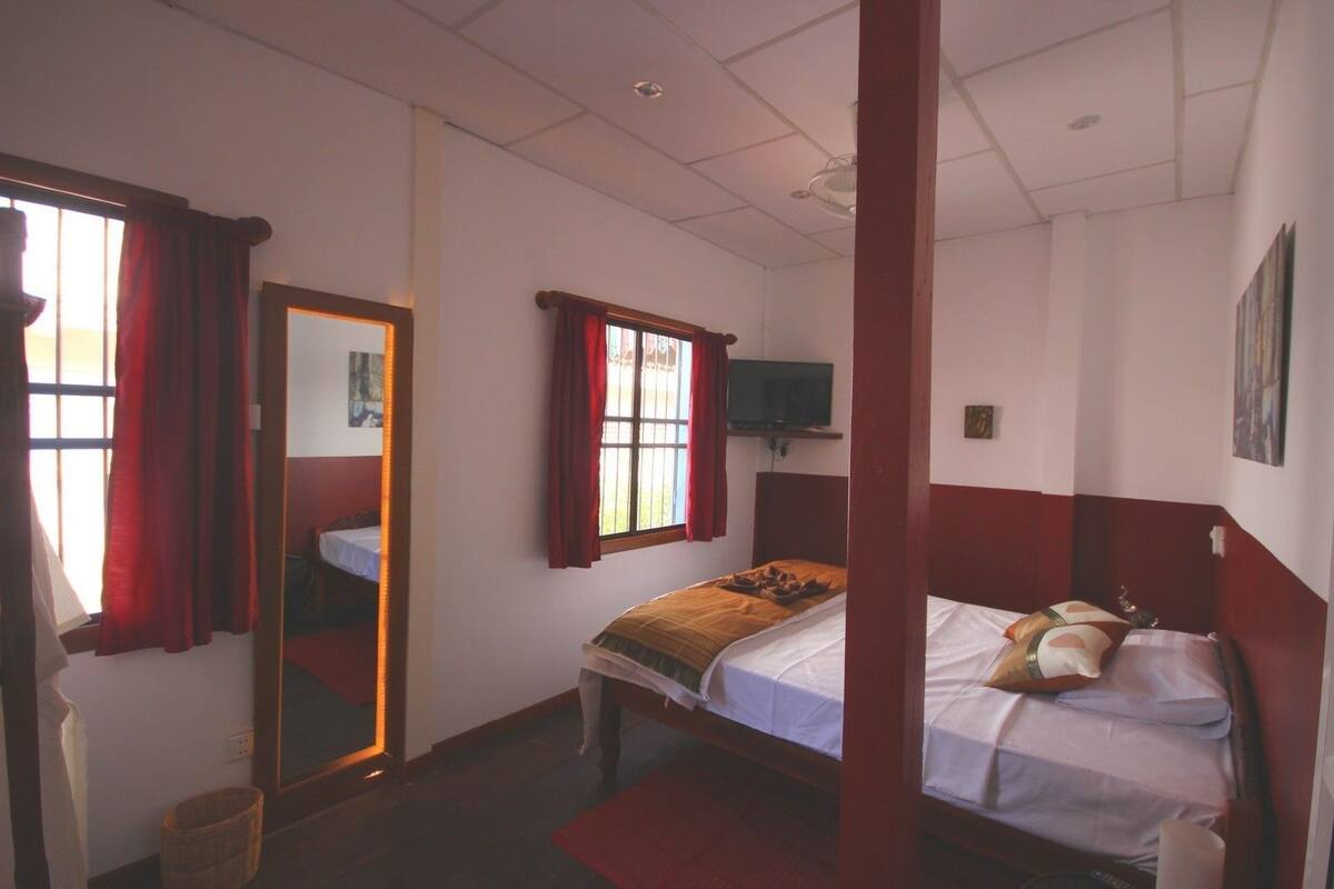Una habitación con cama, espejo, ventana con cortinas, televisor en la pared y una puerta con marco rojo.
