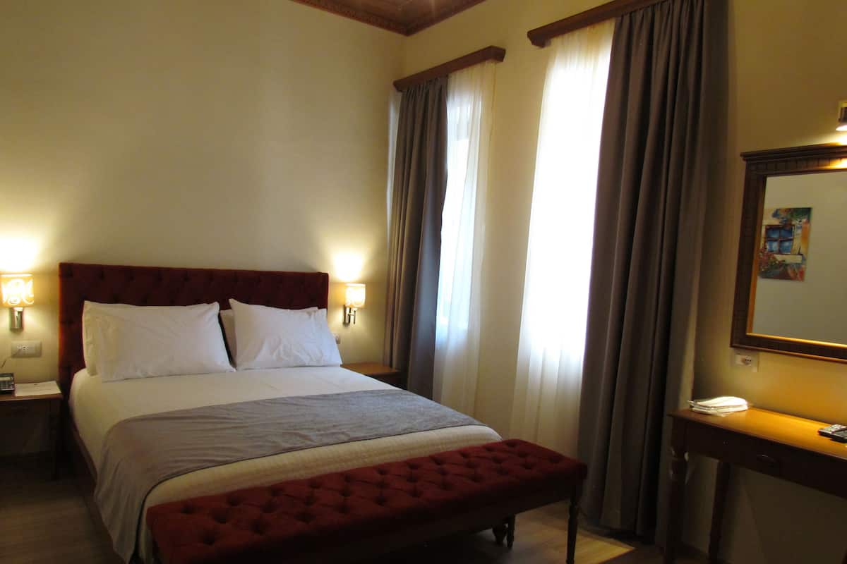 A hotel room with a large bed, a bedside table with a lamp, a desk with a chair, and a window with curtains.
