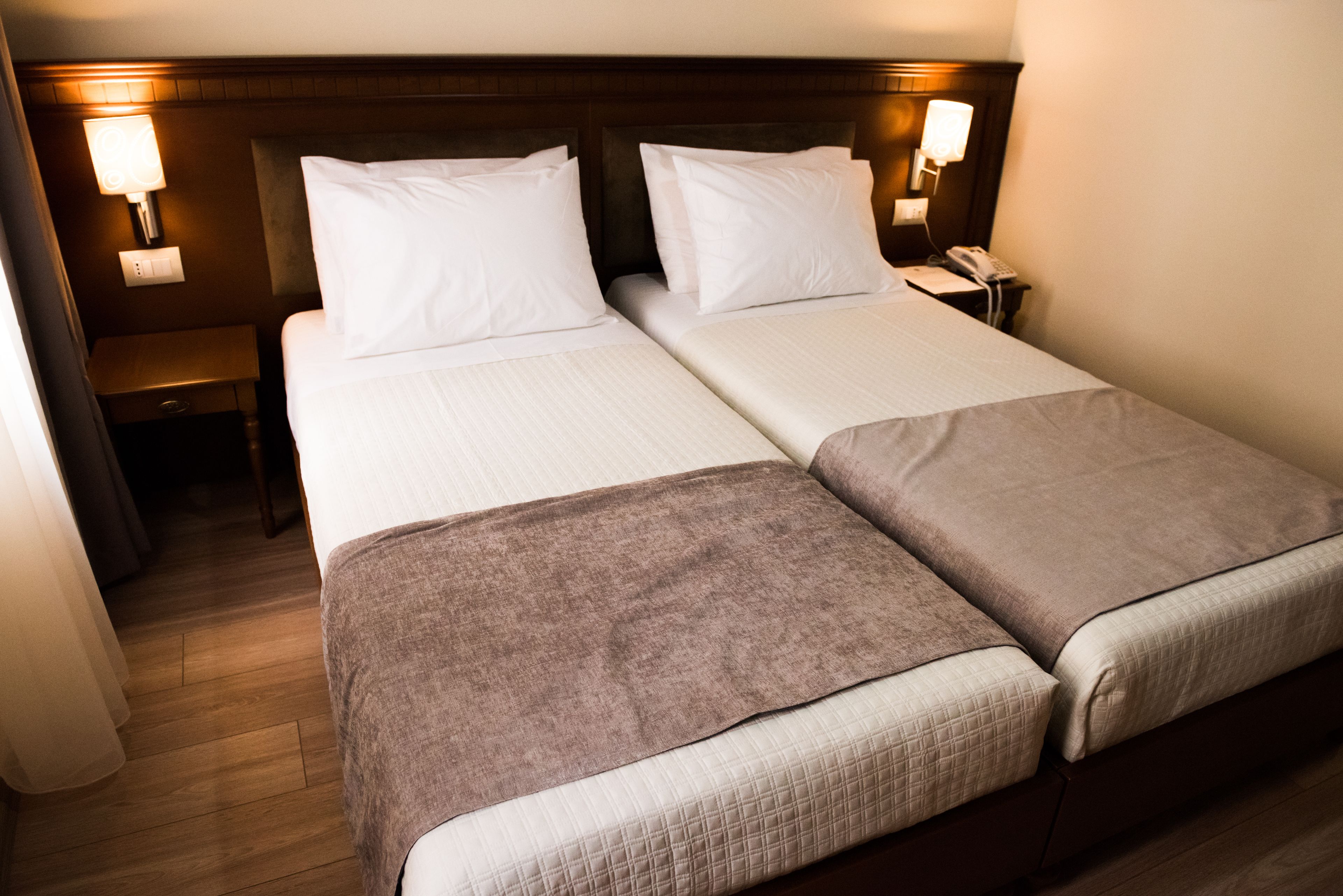 Habitación de hotel con dos camas, cada una con ropa blanca y una manta marrón. Hay lámparas de noche y un teléfono en la mesita.