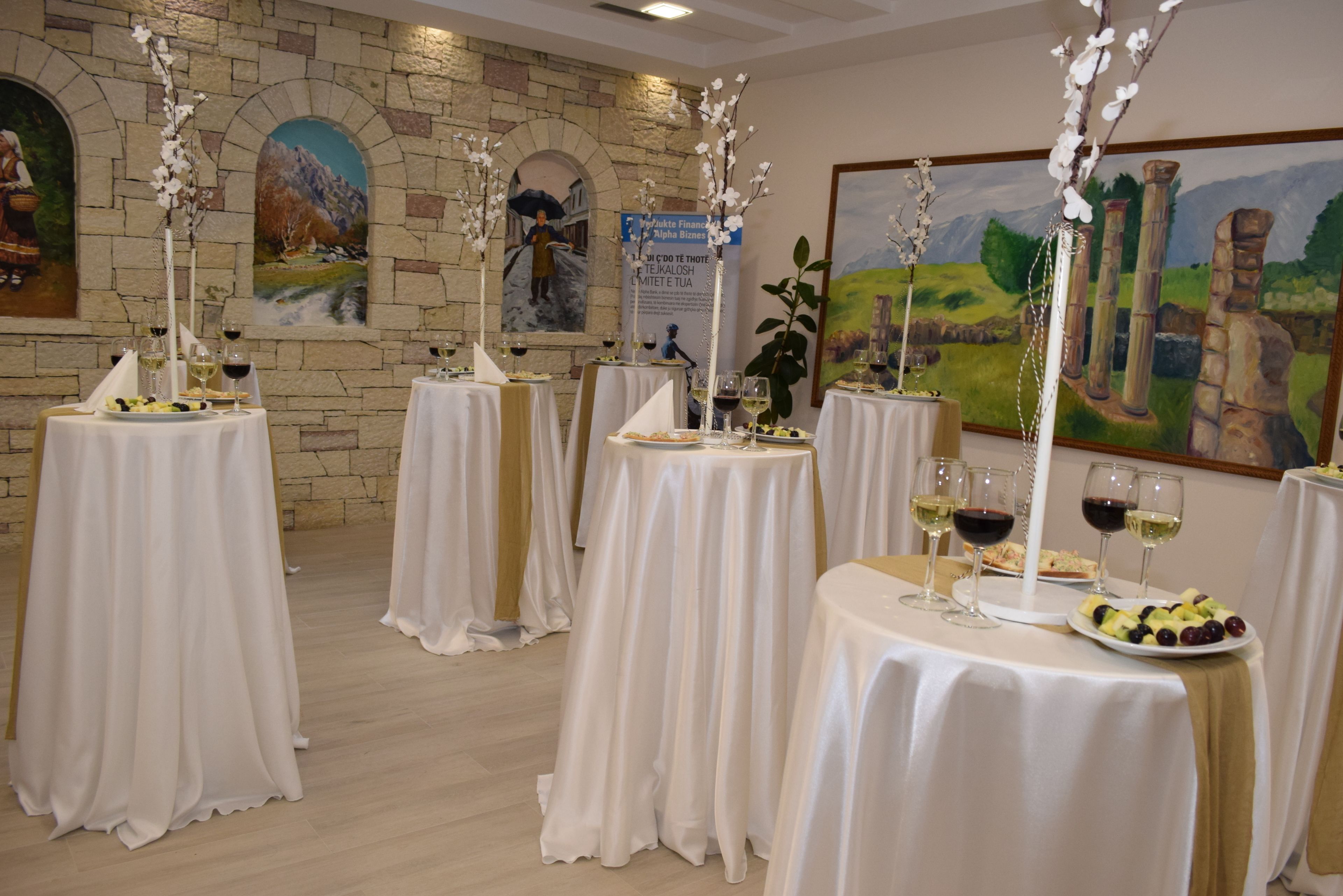 A room with multiple round tables covered with white tablecloths, each set with wine glasses and plates of fruit.