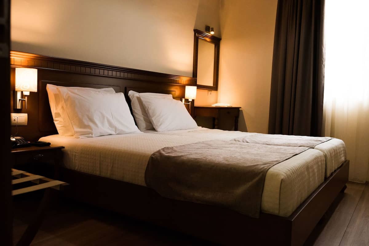 A hotel room with a large bed, bedside tables, a nightstand, a mirror, and a window with curtains.