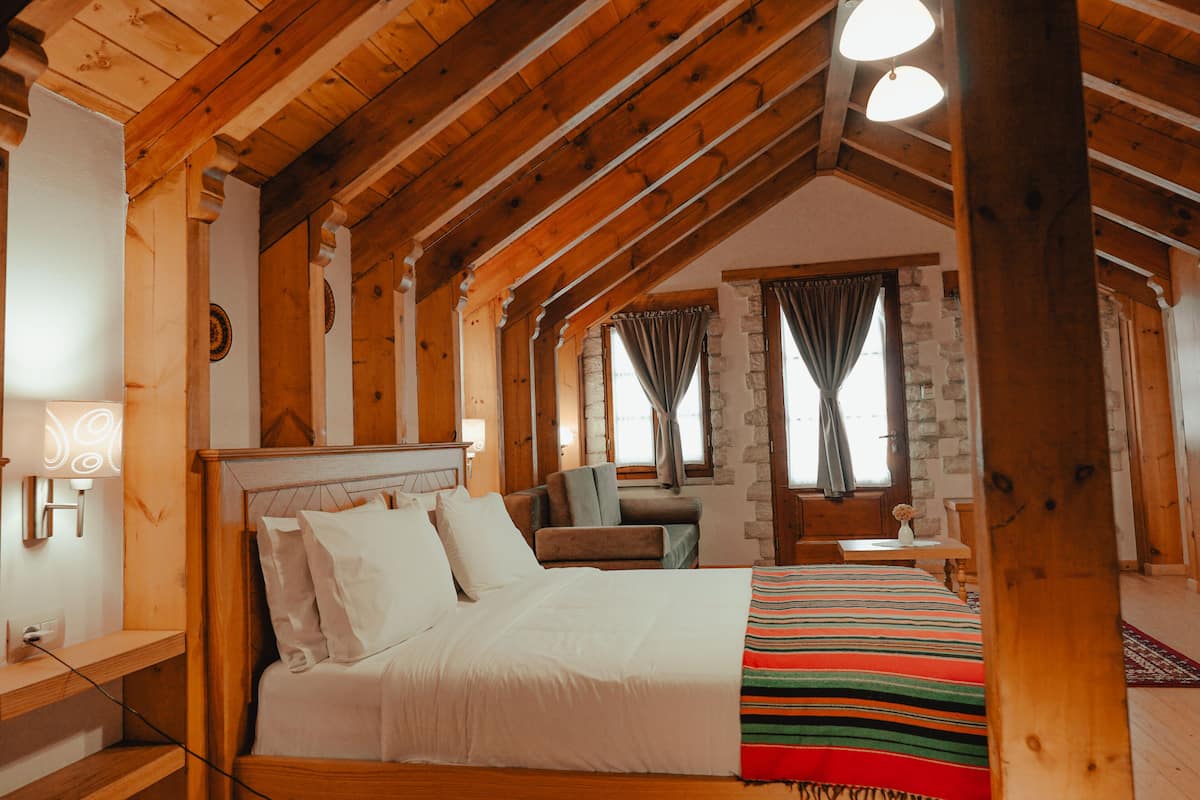 A cozy bedroom with a wooden ceiling, a bed with white bedding, a striped throw blanket, and a stone wall.