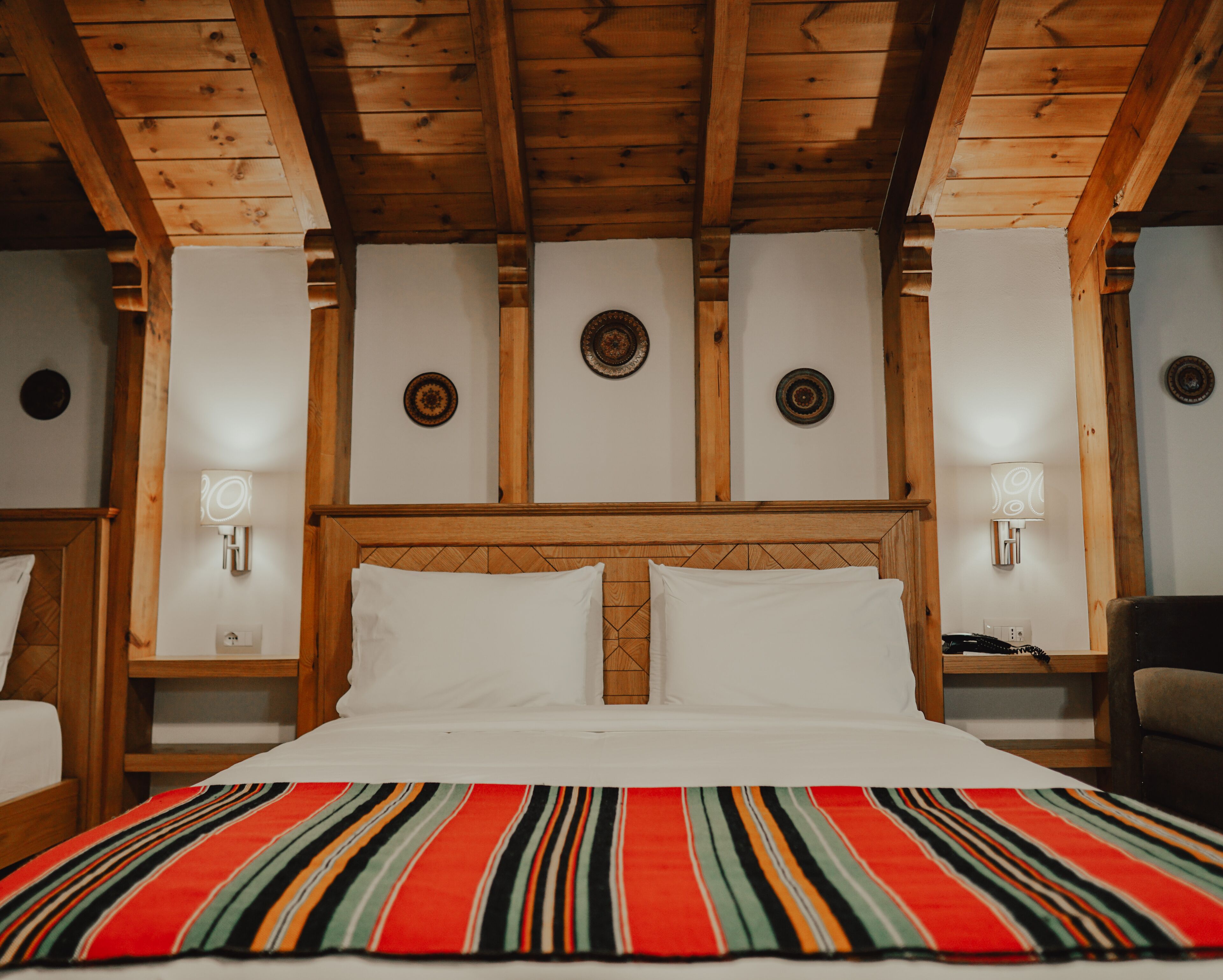 Habitación de hotel con techo de madera, cama grande con ropa blanca, manta a rayas de colores y lámparas fijadas a la pared.