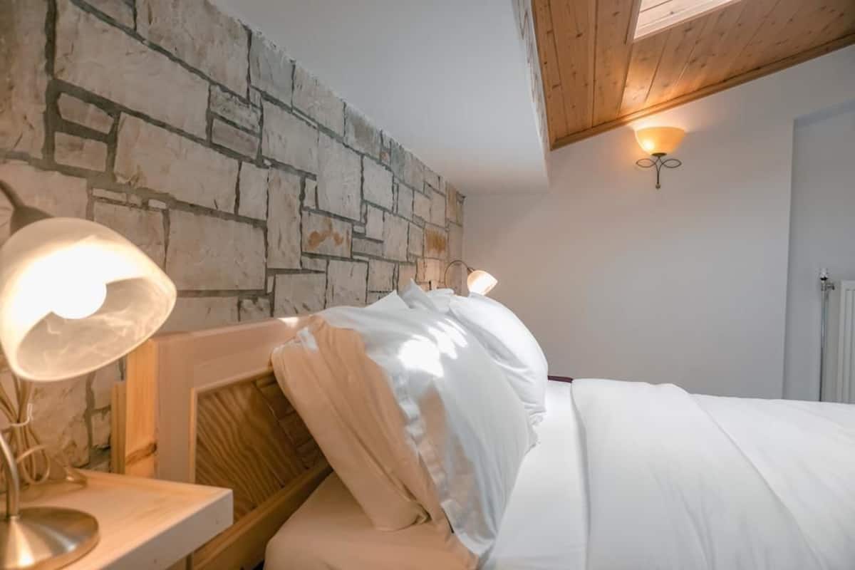 A bedroom with a stone wall, a wooden headboard, a bedside lamp, and a bed with white bedding.