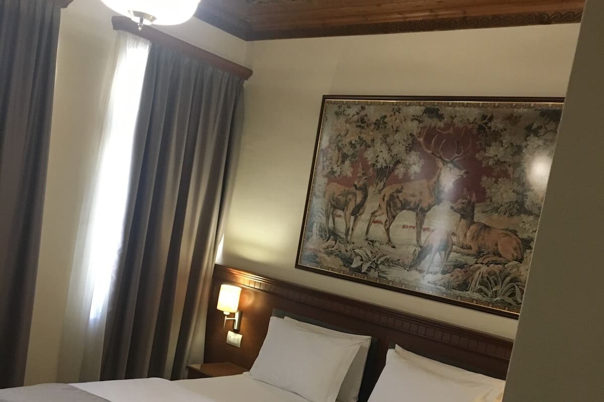 A hotel room with a bed, a headboard, a wall-mounted painting, a window with curtains, and a ceiling light.
