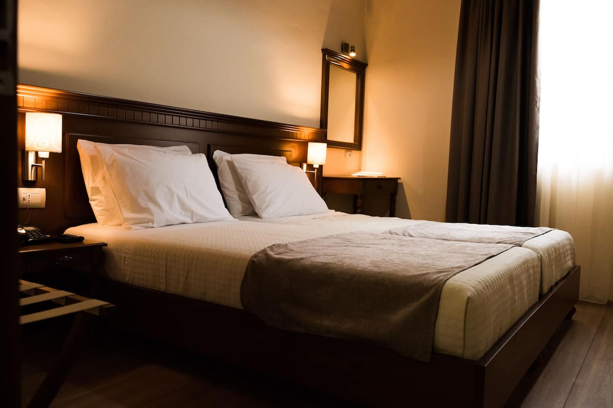 A hotel room with a large bed, a nightstand with a lamp, a mirror, and a window with curtains.