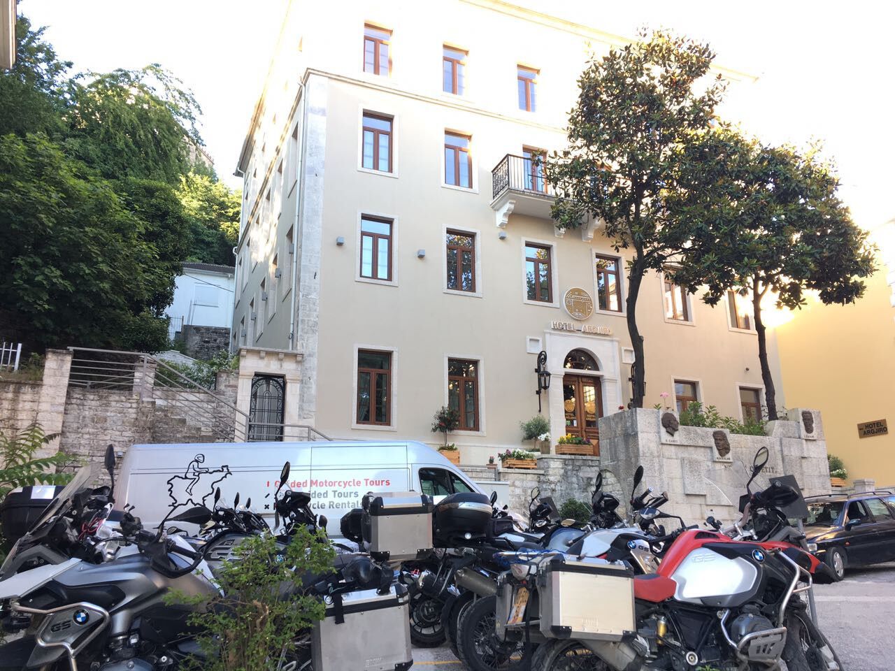 Hotellissa on useita kerroksia, valkoinen julkisivu ja parveke. Sen edessä on parkkeerattuna useita moottoripyöriä, ja lähellä on pakettiauto, jossa on kyltti, joka mainostaa moottoripyöräretkiä.