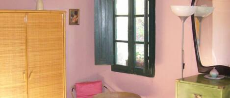 Ein Zimmer mit einem Bett, einem grünen Schminktisch, einem Stuhl, einem kleinen Tisch, einem Fenster mit Rollläden und einer Vase mit Blumen.
