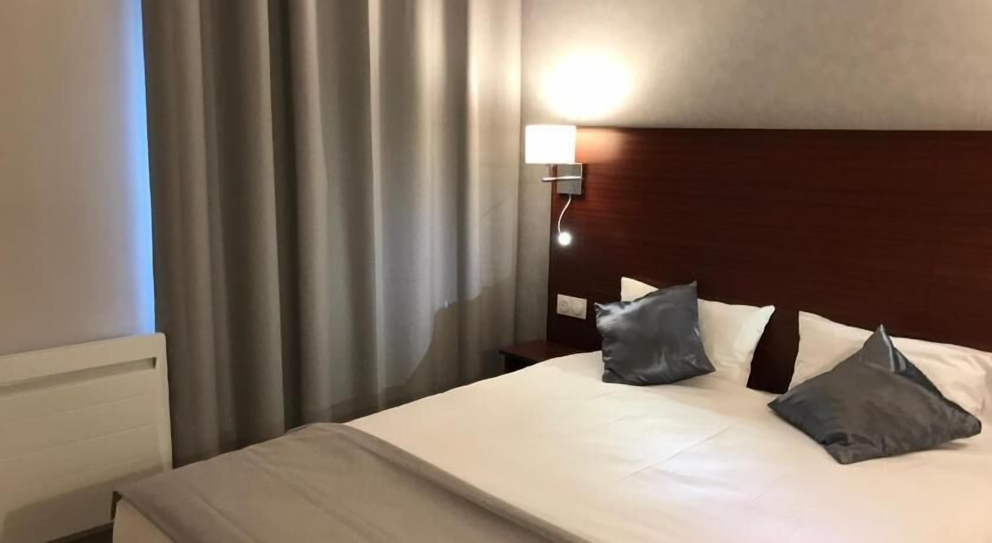 A hotel room with a bed, curtains, a nightstand, and a wall-mounted lamp.
