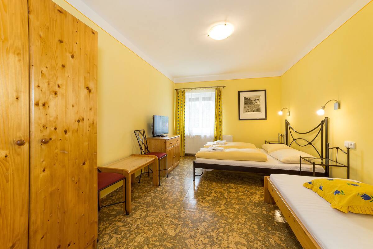 A room with a wooden wardrobe, a desk with a chair, a bed with a yellow blanket, a bedside table, and a window with curtains.