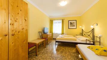 A room with a wooden wardrobe, a desk with a chair, a bed with a yellow blanket, a bedside table, and a window with curtains.