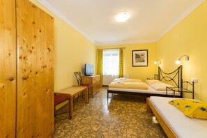 A room with a wooden wardrobe, a desk with a chair, a bed with a yellow blanket, a bedside table, and a window with curtains. - Villa Gardena (Cesky Krumlov)