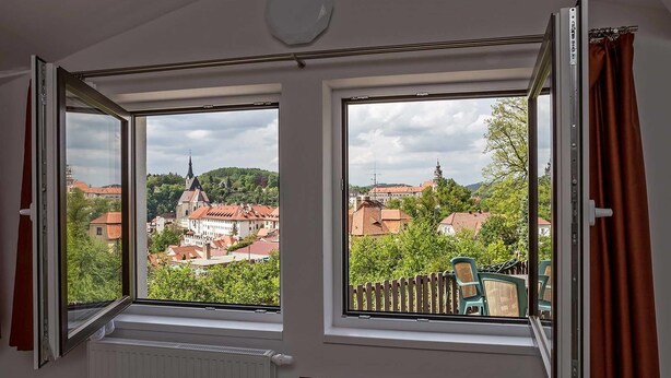A room with two open windows offering a view of a town with red-roofed buildings and a church steeple. - Villa Gardena (Cesky Krumlov)