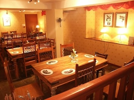 Un área de comedor con mesas y sillas de madera, una pared decorativa con cuadros enmarcados, y una escalera con barandilla de madera.