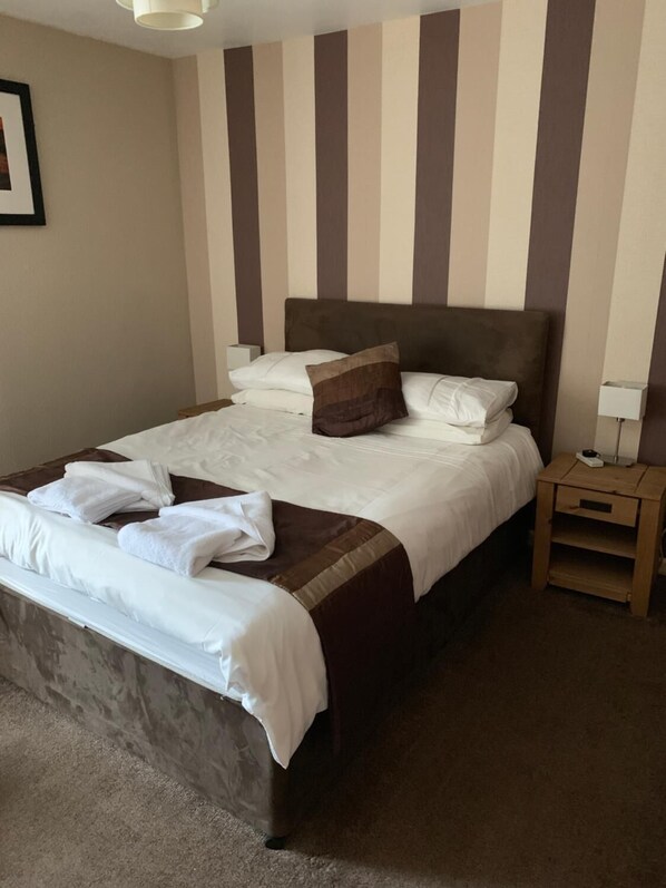 A neatly made bed with white linens and a brown pillow, a wooden nightstand with a lamp, and a striped wall in the background. - The White Swan Hotel (Duns)