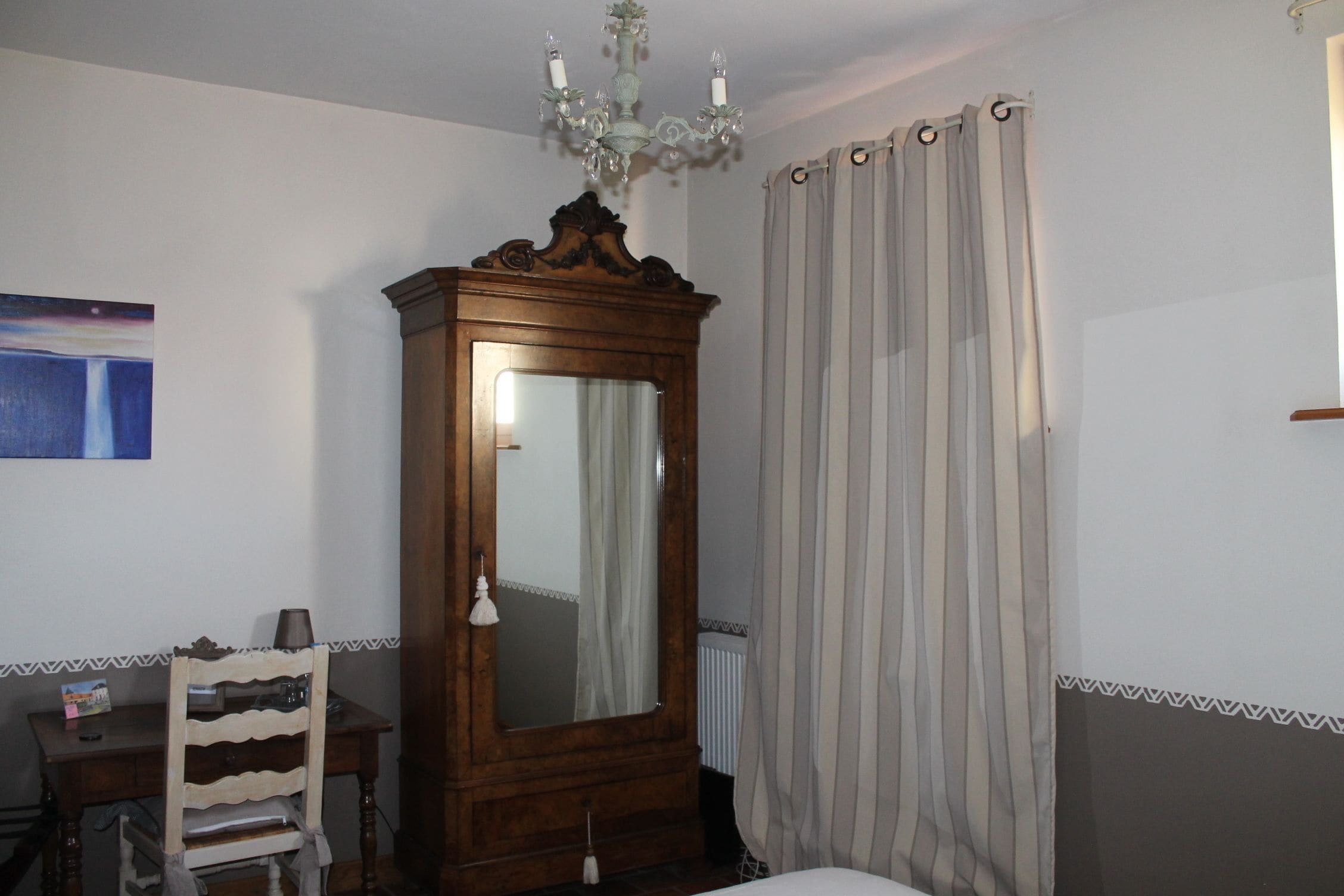 Ein Schlafzimmer mit einem hölzernen Kleiderschrank, einem Sessel, einem Bett, einem Spiegel, einer Chandelier und einem Fenster mit Vorhängen.