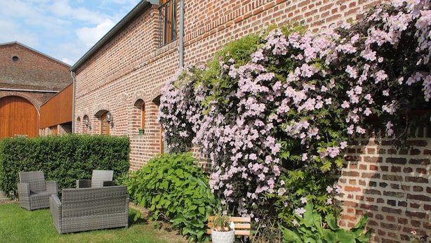 Een bakstenen gebouw met een tuin, voorzien van een tafel, stoelen, een bankje en bloeiende planten.