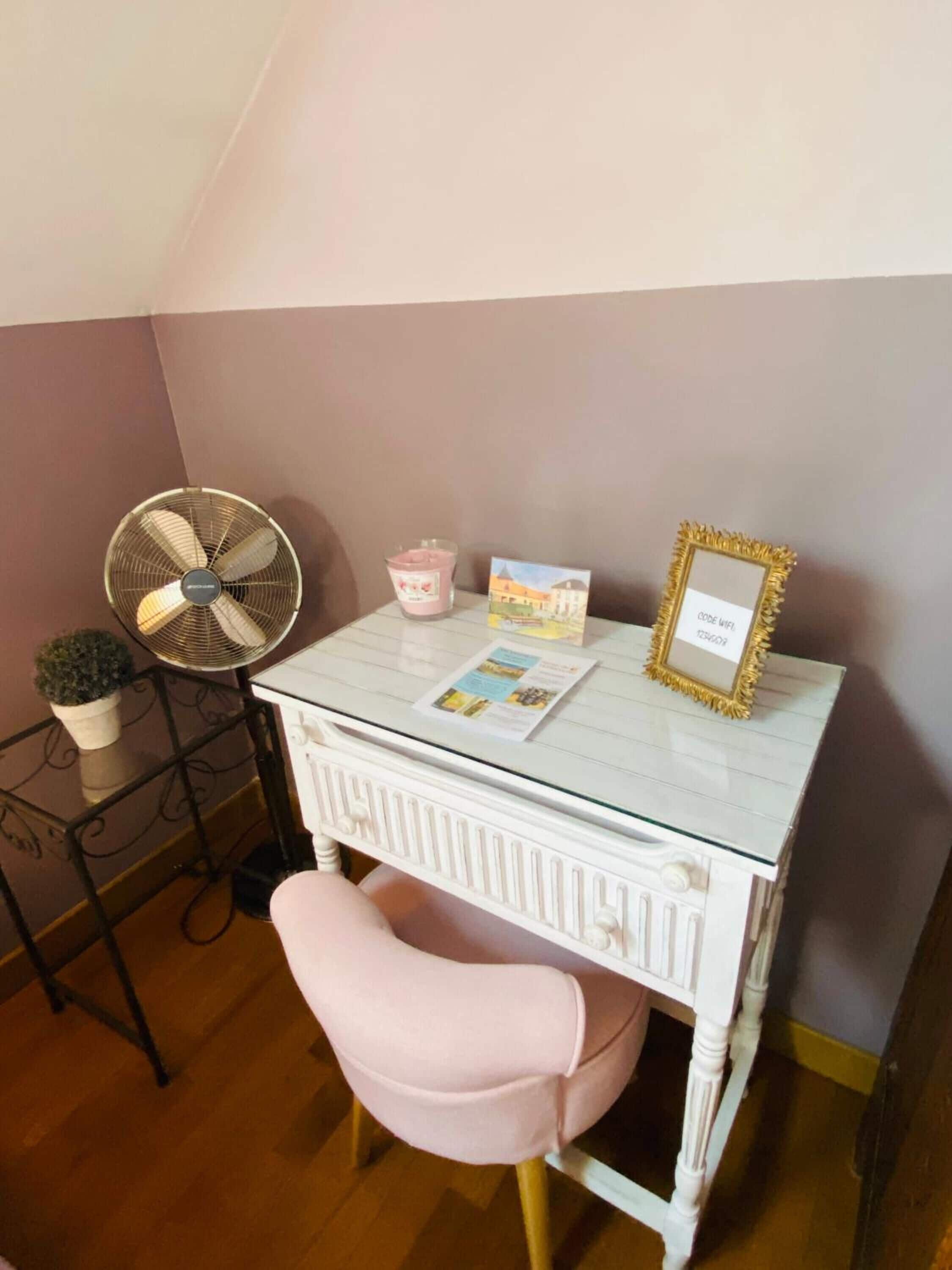 Un pequeño escritorio blanco con una silla rosa, un ventilador, una vela y un cuadro en la pared.
