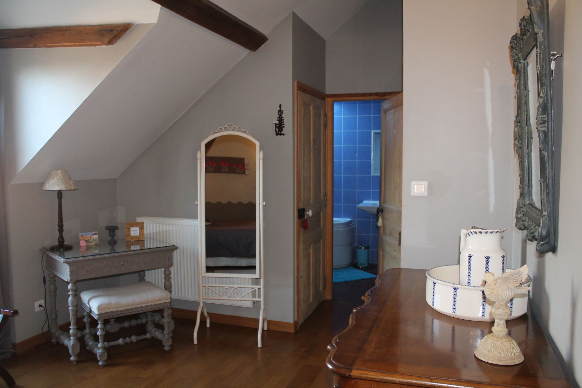 Una stanza con soffitto spiovente, una scrivania in legno con lampada, una panca bianca, un uccellino decorativo su un tavolo e vista su un bagno con piastrelle blu.