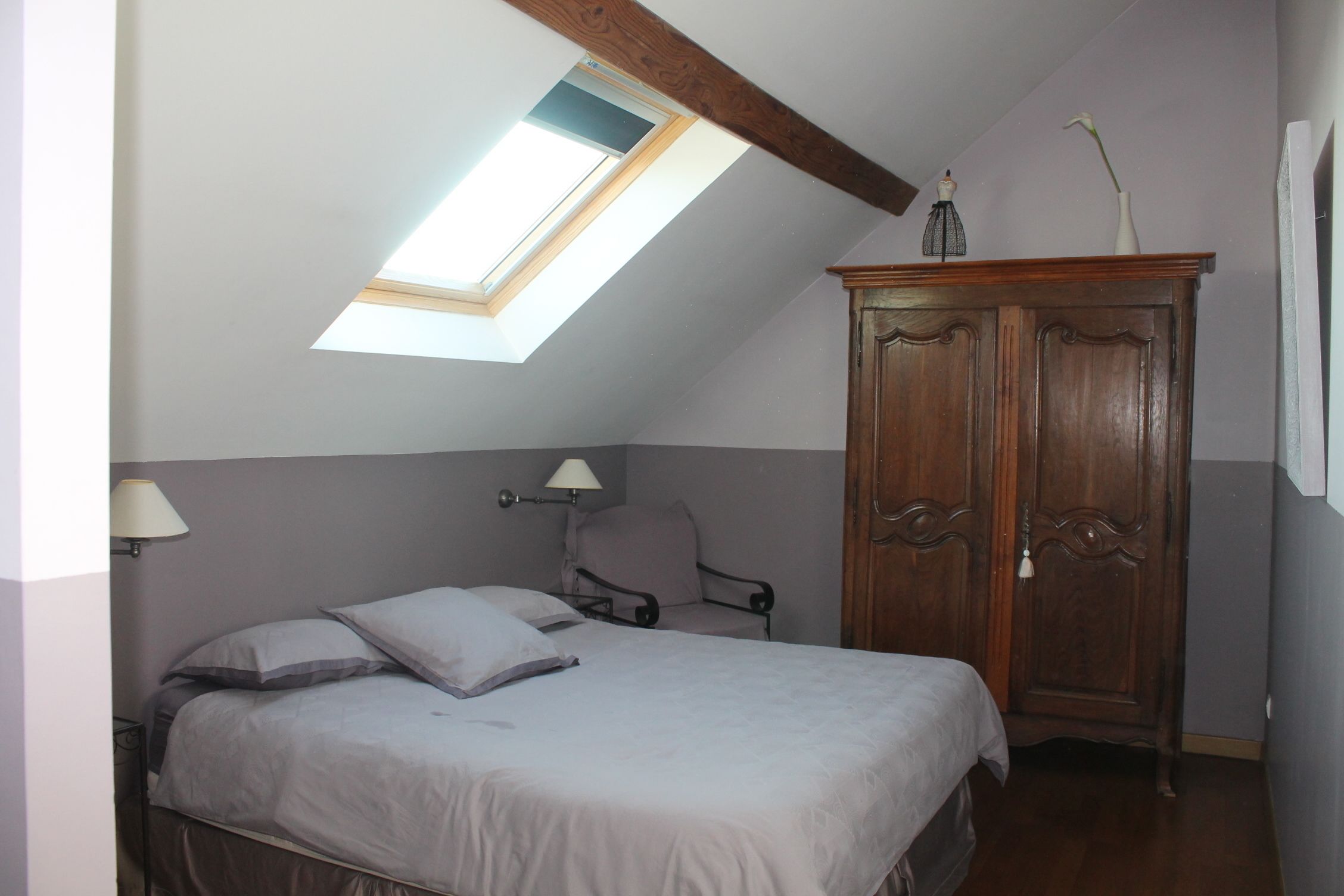 A bedroom with a sloped ceiling, a bed, a chair, a wardrobe, and a skylight.