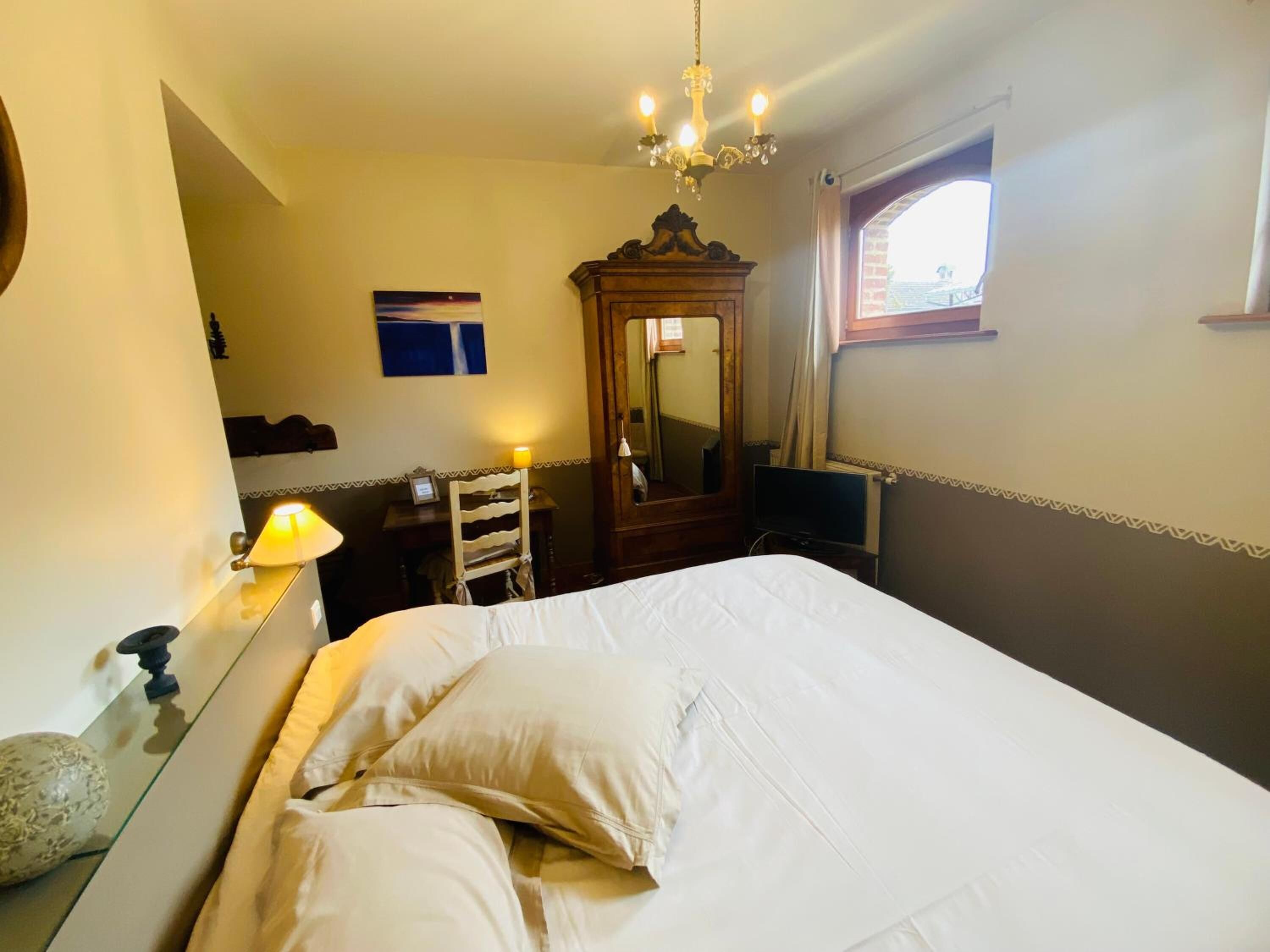 A hotel room with a bed, a wooden dresser, a mirror, a desk with a lamp, and a window with curtains.