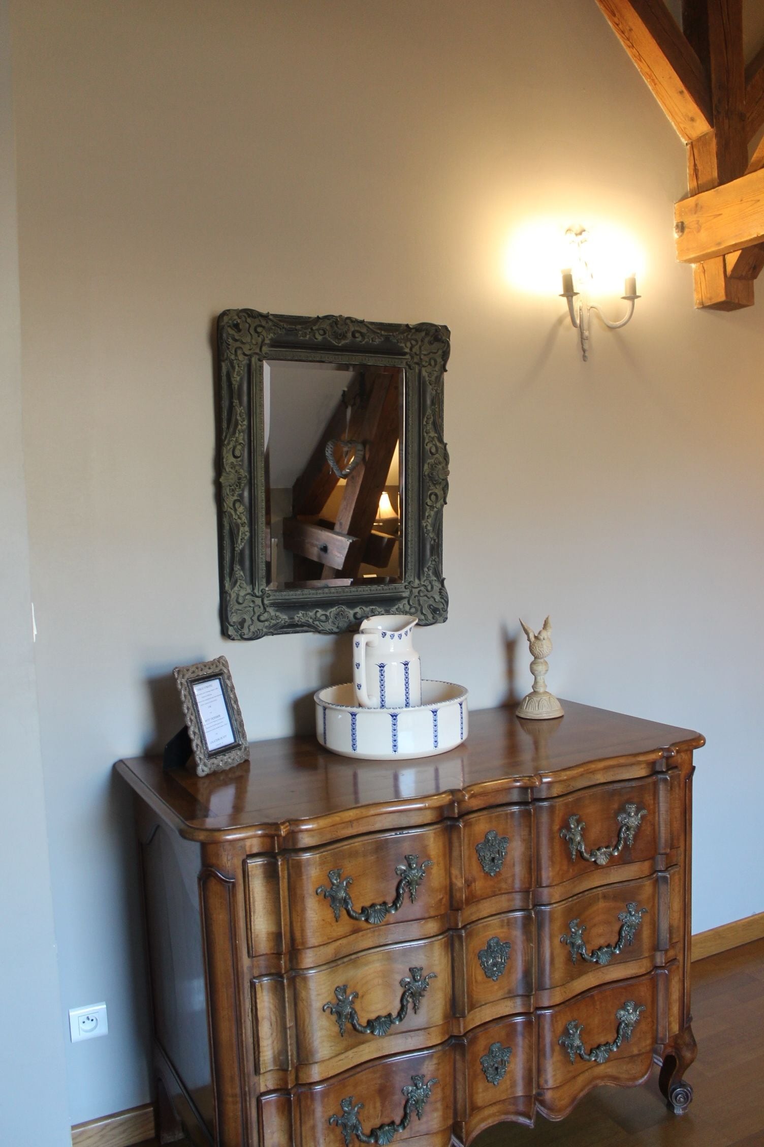 A wooden dresser with ornate handles, a mirror above it, and a wall-mounted light fixture.