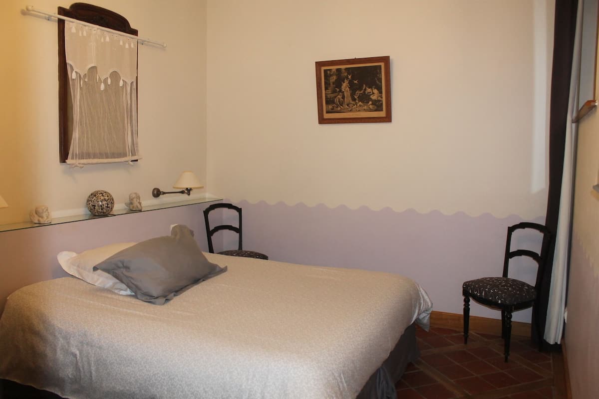 Een slaapkamer met een bed, twee stoelen, een tafeltje met een lamp, een lijst met een afbeelding aan de muur, en een raam met gordijn.