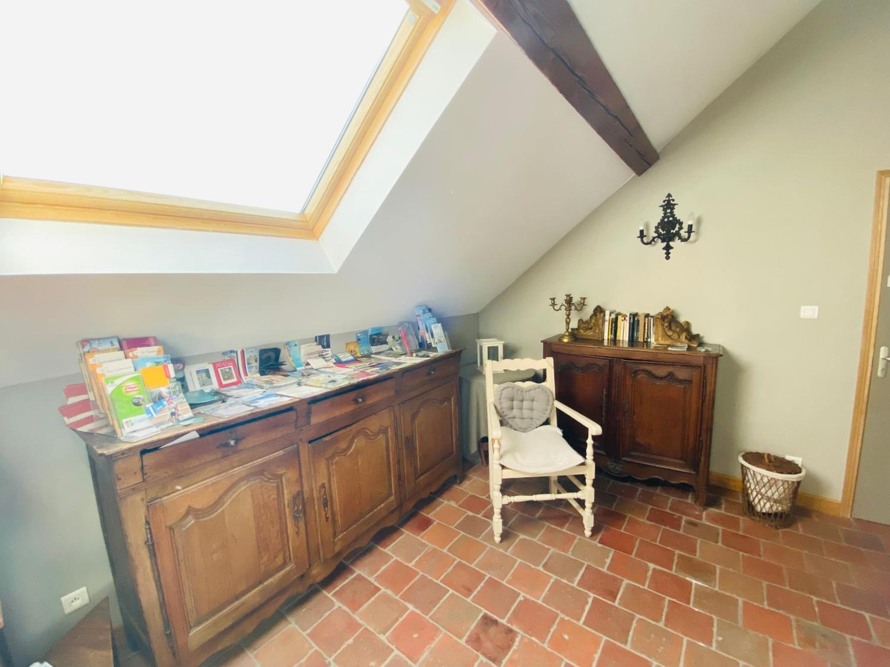 Una habitación con techo inclinado, tragaluz, escritorio de madera con libros, una silla blanca y un mueble pequeño.