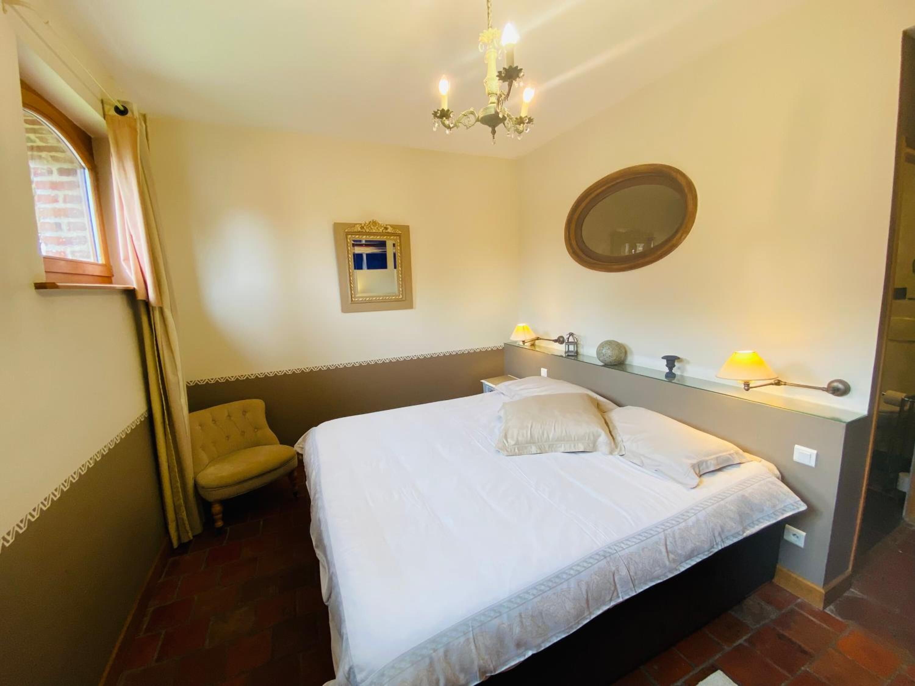 A bedroom with a bed, a chair, a mirror, and a chandelier.