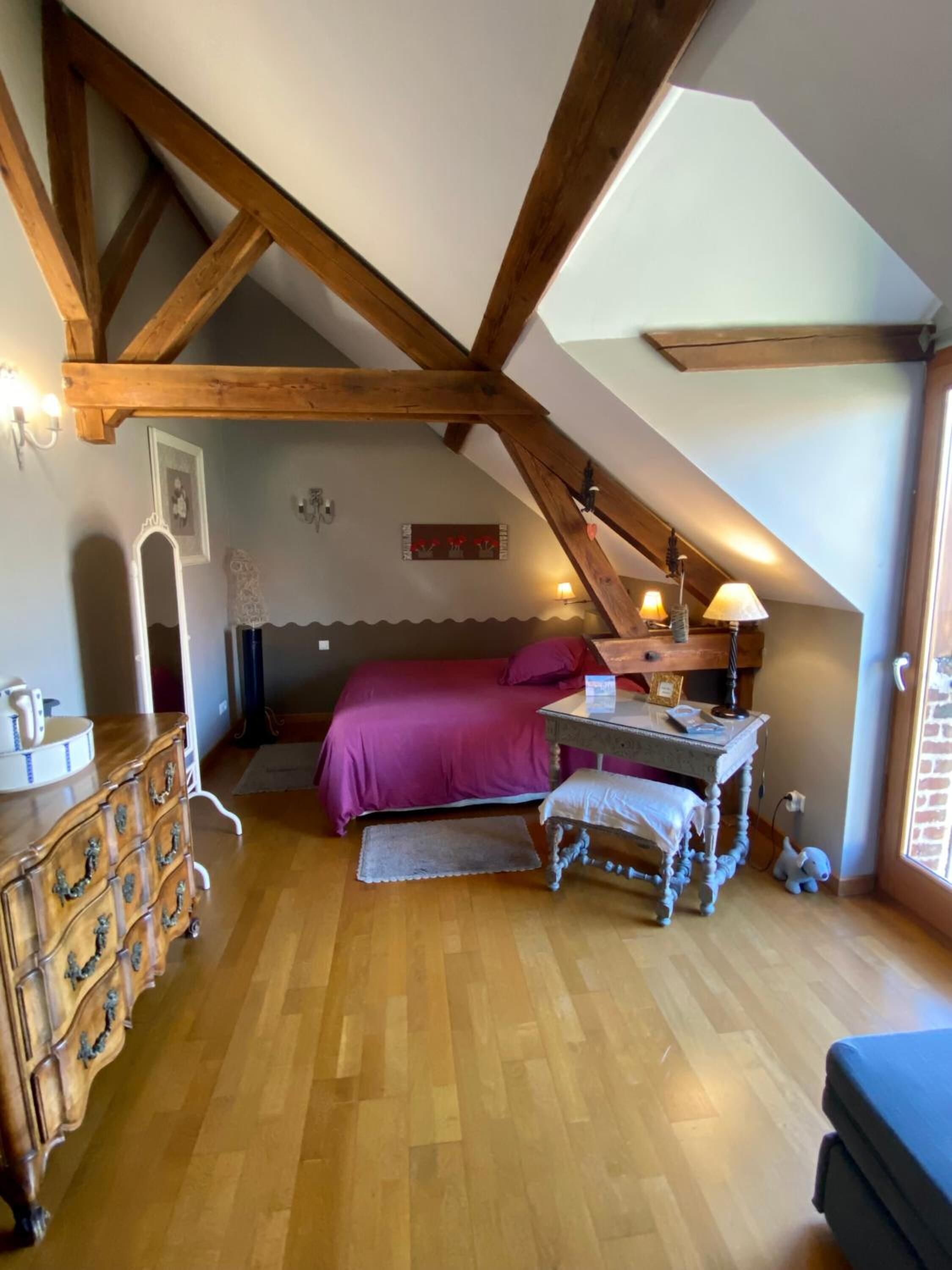 A bedroom with a sloped ceiling, wooden beams, a bed with a purple bedspread, a dresser, a desk with a lamp, and a small rug.