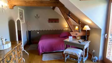 Ein Schlafzimmer mit schrägem Dach, Holzbalken, einem Bett mit violettem Überwurf, einem Schminktisch, einem Schreibtisch mit Lampe und einem kleinen Teppich.