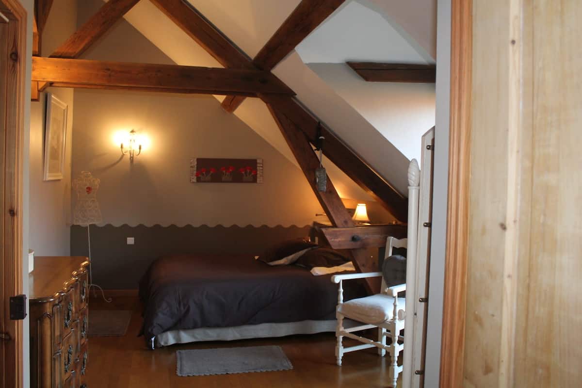 Een slaapkamer met een schuine kap, houten balken, een bed, een stoel, een lamp en een wandlamp.